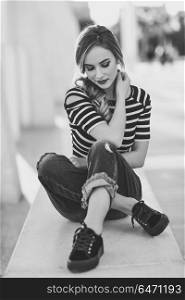 Blonde woman, model of fashion, sitting in urban background.. Blonde woman, model of fashion, sitting on a bench in urban background with eyes closed. Thoughtful young girl wearing striped t-shirt and blue jeans in the street. Pretty russian female with pigtail.