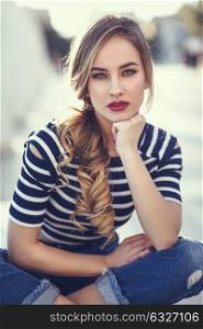 Blonde woman, model of fashion, sitting on a bench in urban background. Beautiful young girl wearing striped t-shirt and blue jeans in the street. Pretty russian female with pigtail.