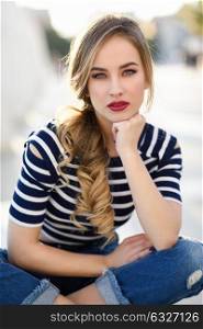 Blonde woman, model of fashion, sitting on a bench in urban background. Beautiful young girl wearing striped t-shirt and blue jeans in the street. Pretty russian female with pigtail.