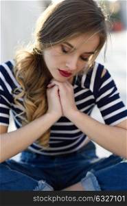 Blonde woman, model of fashion, sitting on a bench in urban background with eyes closed. Thoughtful young girl wearing striped t-shirt and blue jeans in the street. Pretty russian female with pigtail.