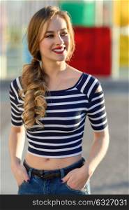 Blonde woman, model of fashion, smiling in urban background. Beautiful young girl wearing striped t-shirt and blue jeans in the street. Pretty russian female with pigtail.