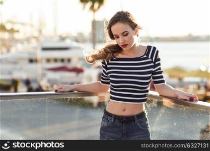 Blonde woman, model of fashion, standing in a harbor with boats and sea in the background. Beautiful young girl wearing striped t-shirt and blue jeans. Pretty russian female with pigtail.
