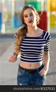 Blonde woman, model of fashion, standing in urban background. Beautiful young girl wearing striped t-shirt and blue jeans in the street. Pretty russian female with pigtail.