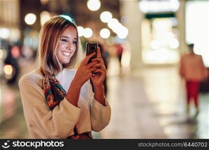 Blonde woman taking photograph with smartphone at night in the street. Defocused city lights at the background. Pretty girl with pigtail hairstyle at night smiling.