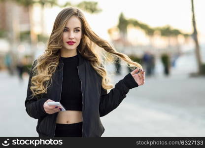 Blonde woman texting with her smart phone in urban background. Beautiful young girl wearing black jacket walking in the street. Pretty russian female with long wavy hair hairstyle.