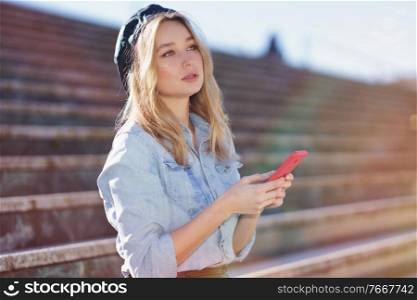 Blonde woman using her smartphone sitting on some city steps, wearing a denim shirt and black beret.. Blonde woman using a smartphone sitting on some city steps, wearing a denim shirt and black beret.