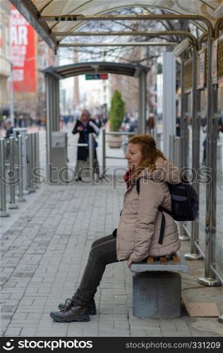 Blonde woman waiting for the tram at the stop.She is tired and is sitting on the bench.