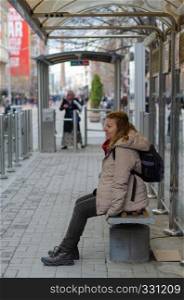 Blonde woman waiting for the tram at the stop.She is tired and is sitting on the bench.