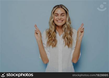 Blonde woman with closed eyes making wish while holding both of her hands with fingers crossed, dreaming about future while standing isolated in front of blue background in studio. Blonde woman with closed eyes making wish