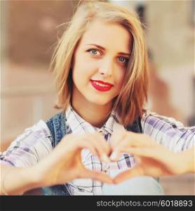 Blonde young hipster girl holding hands in heart shape framing. Portrait of smiling woman having fun outside. Valentines Day. Photo toned style instagram filters