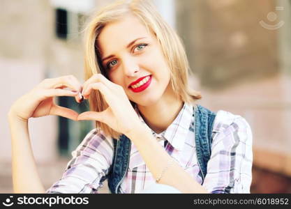 Blonde young hipster girl holding hands in heart shape framing. Portrait of smiling woman having fun outside. Valentines Day. Photo toned style instagram filters