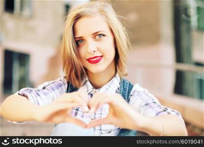 Blonde young hipster girl holding hands in heart shape framing. Portrait of smiling woman having fun outside. Valentines Day. Photo toned style instagram filters