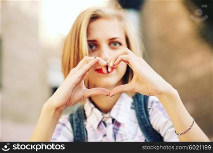 Blonde young hipster girl holding hands in heart shape framing. Portrait of smiling woman having fun outside. Valentines Day.