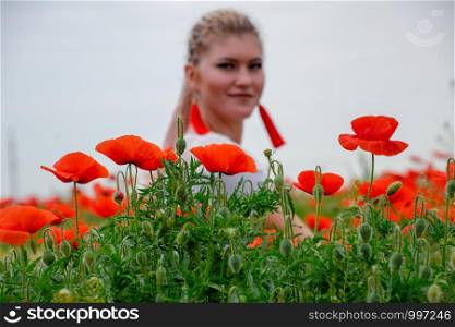 Blonde young woman in a red skirt and white shirt, red earrings is in the middle of a poppy field.. Blonde young woman in red skirt and white shirt, red earrings is in the middle of a poppy field.