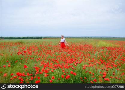 Blonde young woman in a red skirt and white shirt, red earrings is in the middle of a poppy field.. Blonde young woman in red skirt and white shirt, red earrings is in the middle of a poppy field.