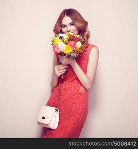 Blonde young woman in elegant red dress. Girl posing on a beige background with handbag. Jewelry and hairstyle. Lady with spring bouquet of flowers. Fashion photo