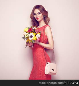 Blonde young woman in elegant red dress. Girl posing on a beige background with handbag. Jewelry and hairstyle. Lady with spring bouquet of flowers. Fashion photo