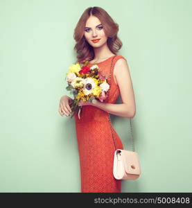 Blonde young woman in elegant red dress. Girl posing on a green background with handbag. Jewelry and hairstyle. Lady with spring bouquet of flowers. Fashion photo