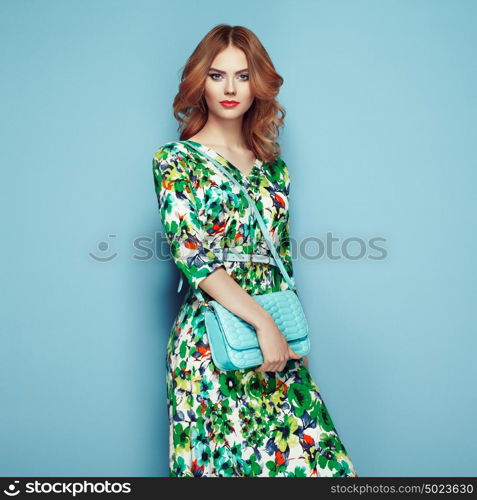 Blonde young woman in floral spring summer dress. Girl posing on a pink background. Summer floral outfit. Stylish wavy hairstyle. Fashion photo. Glamour lady with handbag