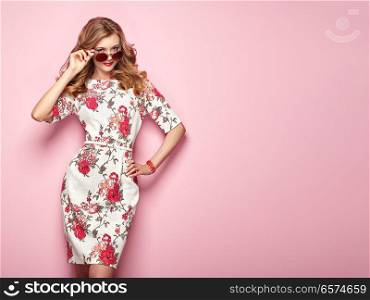 Blonde young woman in floral spring summer dress. Girl posing on a pink background. Summer floral outfit. Stylish wavy hairstyle. Fashion photo. Glamour lady in stylish sunglasses