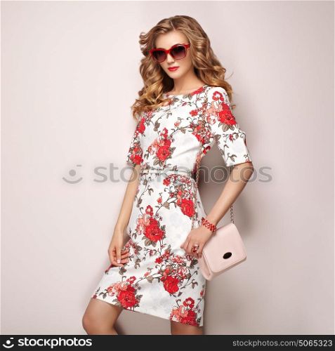 Blonde young woman in floral spring summer dress. Girl posing on a white background. Summer floral outfit. Stylish wavy hairstyle. Fashion photo. Glamour lady in sunglasses with handbag