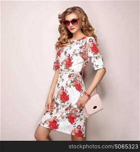 Blonde young woman in floral spring summer dress. Girl posing on a white background. Summer floral outfit. Stylish wavy hairstyle. Fashion photo. Glamour lady in sunglasses with handbag