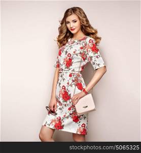 Blonde young woman in floral spring summer dress. Girl posing on a white background. Summer floral outfit. Stylish wavy hairstyle. Fashion photo. Glamour lady with sunglasses and handbag