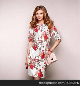 Blonde young woman in floral spring summer dress. Girl posing on a white background. Summer floral outfit. Stylish wavy hairstyle. Fashion photo. Glamour lady with sunglasses and handbag