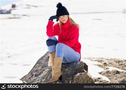 Blonde young woman sitting on a rock in the snowy mountains in winter, in Sierra Nevada, Granada, Spain. Female wearing winter clothes.. Blonde woman sitting on a rock in the snowy mountains in winter, in Sierra Nevada, Granada, Spain.