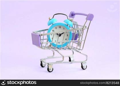 Blue alarm clock in supermarket trolley on violet background. Time resource acquisition concept. Time to sale. Blue alarm clock in supermarket trolley on violet background