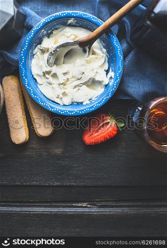 Blue bowl with mascarpone cream and wooden cooking spoon on dark wooden background, top view. Tiramisu preparation