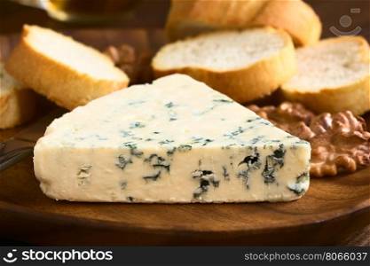 Blue cheese with walnuts and bread slices on wooden plate, photographed with natural light (Selective Focus, Focus on the front of the cheese)