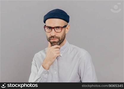 Blue eyed male adult holds chin and looks attentively at camera, ponders on way out or solution, wears spectacles, headgear and formal shirt, models over grey studio wall. Businessman poses indoor