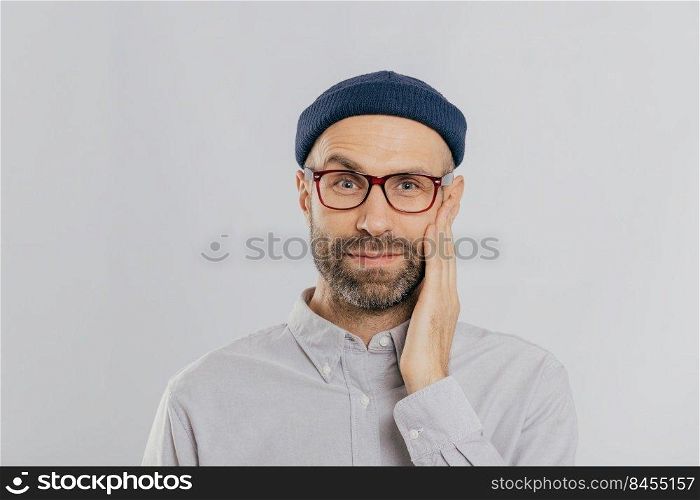 Blue eyed unshaven man raises eyebrow, keeps hand on cheek, looks happily, wears eyewear, dressed in black hat and shirt, expresses positive emotions, isolated over white wall. Facial expressions