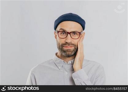Blue eyed unshaven man raises eyebrow, keeps hand on cheek, looks happily, wears eyewear, dressed in black hat and shirt, expresses positive emotions, isolated over white wall. Facial expressions