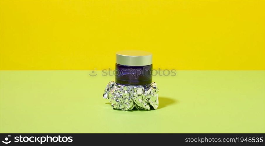 blue glass jar with gray plastic lid for cosmetics on an abstract background with crumpled foil. Branding of cosmetic products cream, mask, serum