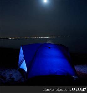 Blue illuminated tent at night on the beach. Moon, stars and reflection in water