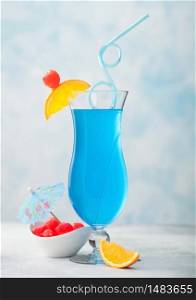 Blue lagoon summer cocktail in classic glass with sweet cocktail cherries and orange slice with umbrella on blue table background.
