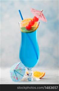 Blue lagoon summer cocktail in classic glass with sweet cocktail cherries and orange slice with umbrella on blue table background.