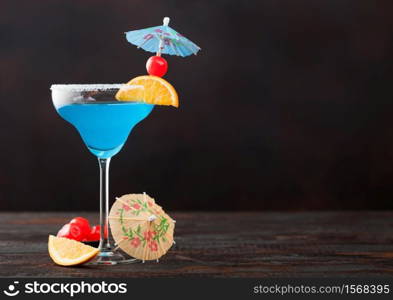 Blue lagoon summer cocktail in margarita glass with sweet cocktail cherries and orange slice with umbrella on dark table background. Space for text