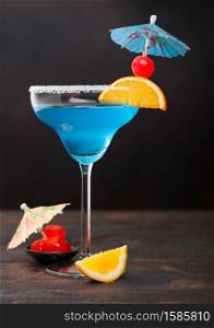Blue lagoon summer cocktail in margarita glass with sweet cocktail cherries and orange slice with umbrella on dark table background.