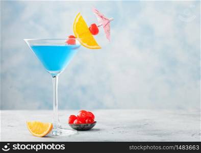 Blue lagoon summer cocktail in martini glass with sweet cocktail cherries and orange slice with umbrella on blue table background. Space for text