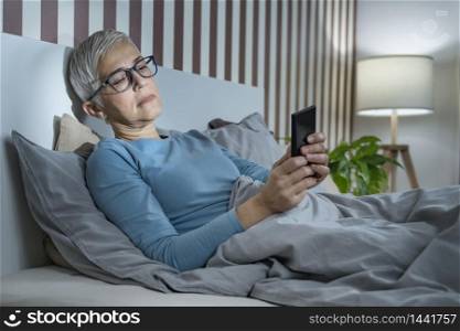 Blue Light Affecting Sleep. Sleepless mature woman suffering from insomnia lying in bed, surfing internet and texting friends via social networks with smartphone,. Blue Light Affecting Sleep