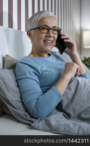 Blue-light blocking glasses. Mature Woman talking over the phone relaxing at home before sleep. Blue-Light Blocking Glasses. Mature Woman Talking over the Phone