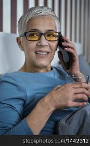 Blue-light blocking glasses. Mature Woman talking over the phone relaxing at home before sleep. Blue-Light Blocking Glasses. Mature Woman Talking over the Phone