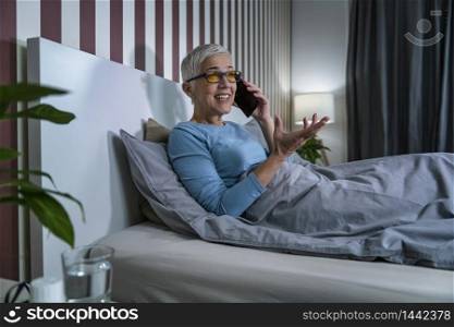 Blue-light blocking glasses. Mature Woman talking over the phone relaxing at home before sleep. Blue-Light Blocking Glasses. Mature Woman Talking over the Phone