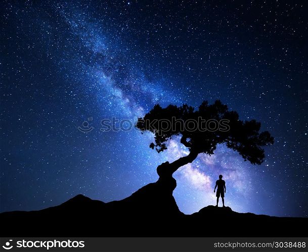 Blue milky way tree and man. Milky Way. Night sky with stars, old tree and silhouette of a standing alone man on the mountain. Blue milky way with light and man. Travel background. Silhouette of a man under the tree. Universe