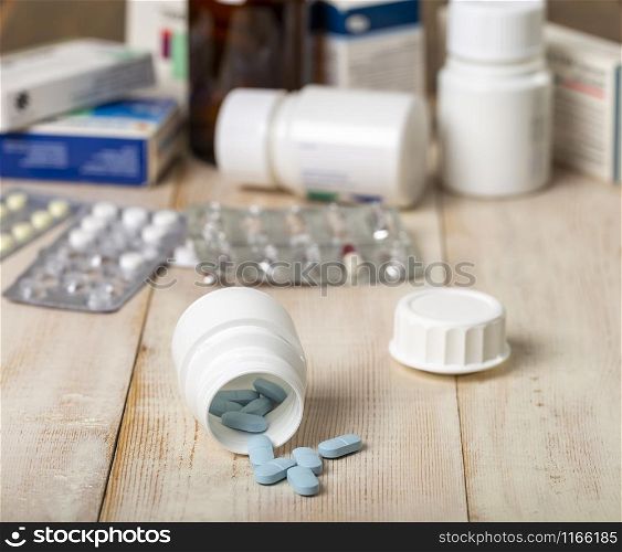 Blue pills spilled from a white plastic bottle. Different tablet boxes and bottles in the background. Bright wooden background. Blue pills spilled from a white plastic bottle. Different drug boxes and bottles in the background.