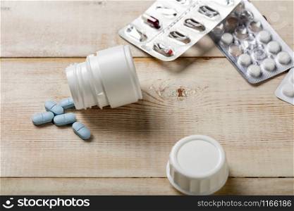 Blue pills spilled from a white plastic bottle. Different tablet boxes and bottles in the background. Bright wooden background. Blue pills spilled from a white plastic bottle. Different drug boxes and bottles in the background.
