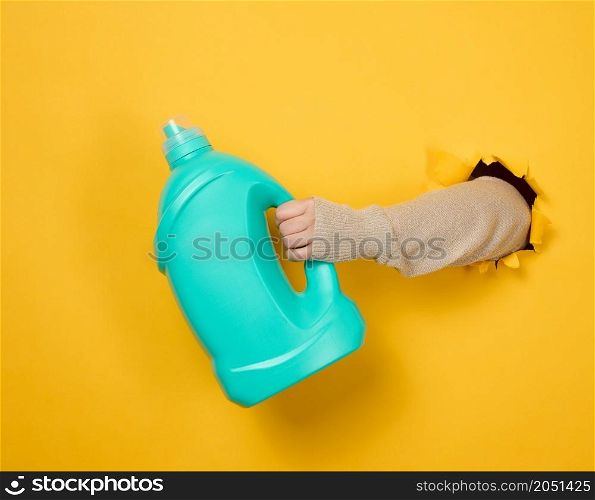 blue plastic bottle with liquid detergent in a female hand on a yellow background. A part of the body sticks out of a torn hole in the background